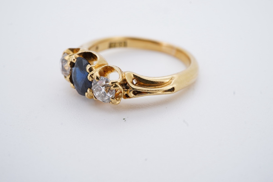 A Victorian sapphire and diamond three-stone ring, late 19th century, set with an oval sapphire between cushion-shaped diamonds totalling approximately 0.30 carats, to a gold mount of scrolled design, size J, maker's mar